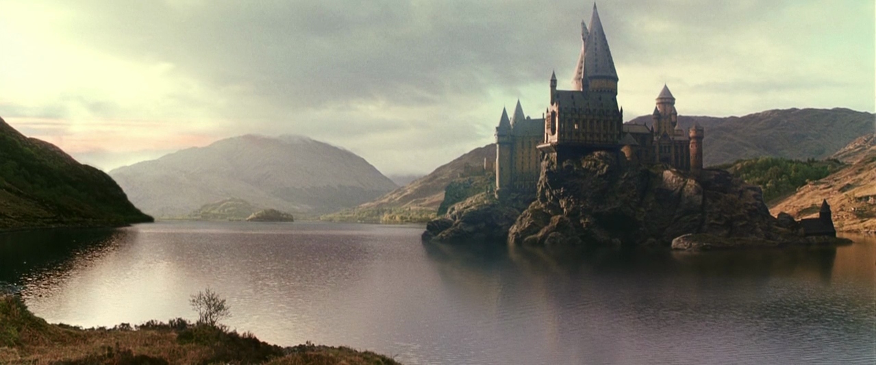 A large stone castle sits perched on a rocky cliff overlooking a calm lake. Mountains rise in the background under a soft overcast sky.