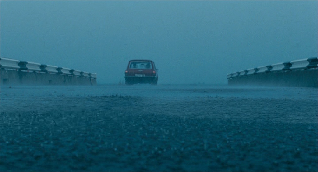 A wide, low-angle shot of a rain-slicked bridge surface during a downpour. A red sedan moves away from the camera into a featureless, grey-blue horizon.