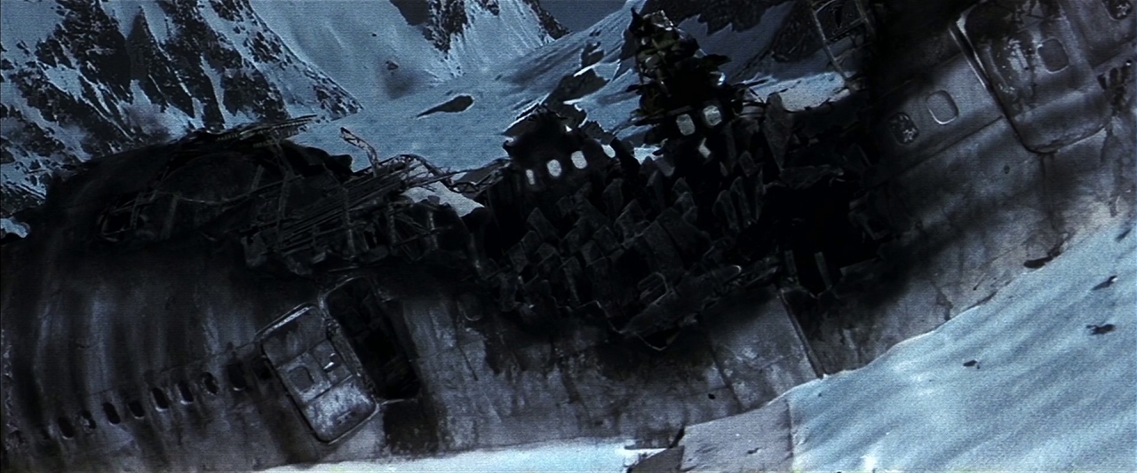 A wreckage of a large commercial aircraft lies partially buried in a snowy mountainside. The mangled fuselage is torn open, revealing interior structural ribs against steep snow-covered peaks.