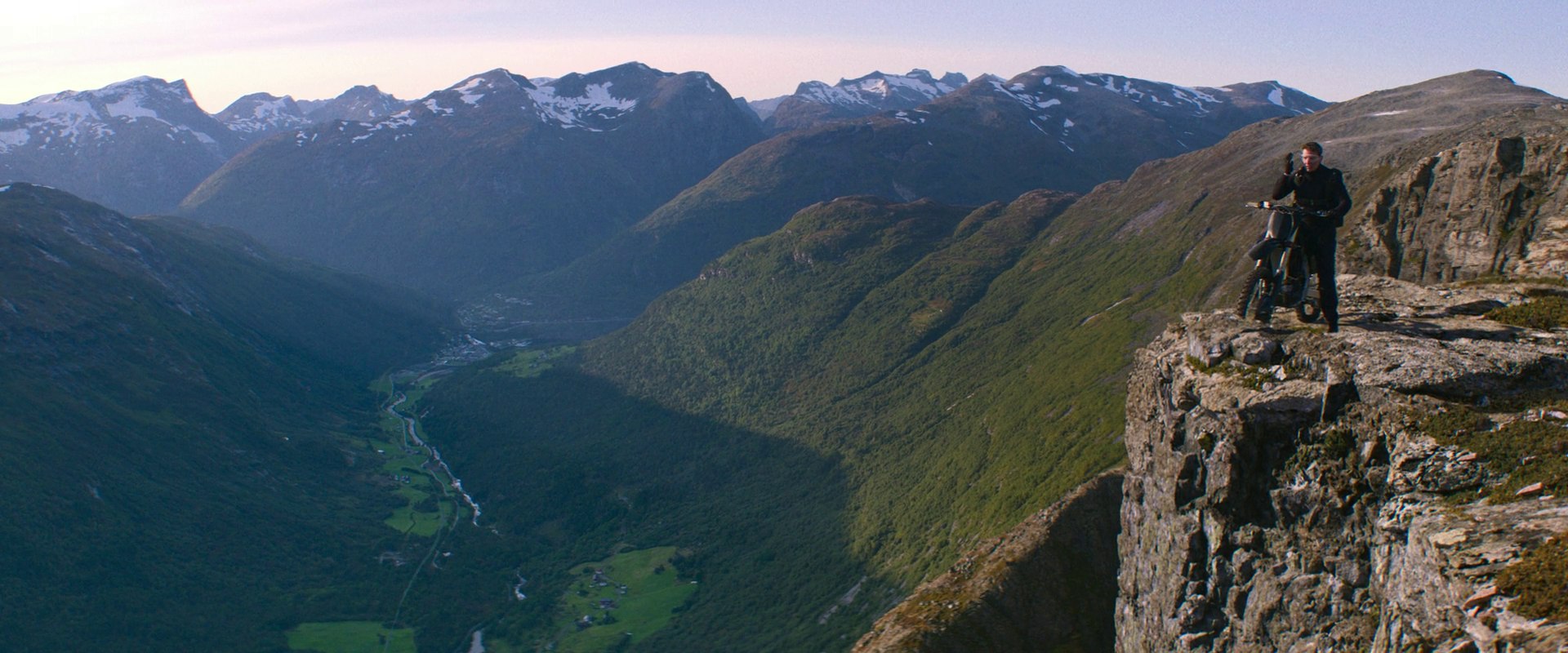 A high mountain peak overlooking a lush, deep valley with snow-capped ridges in distance. A man on a motorcycle sits at the very edge of a sheer cliff drop.