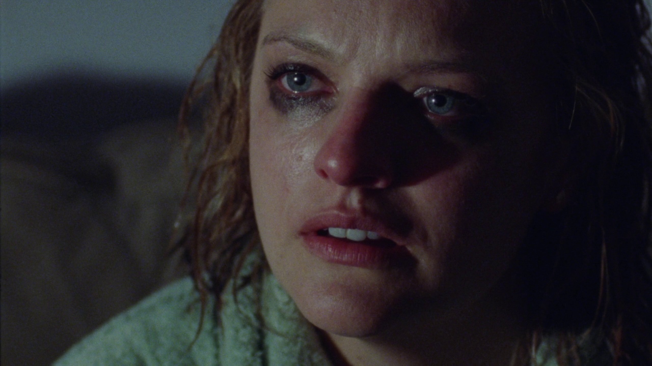 A dimly lit indoor space with dark, indistinct surroundings. A woman with wet hair and smeared makeup stares ahead with a wide-eyed expression.