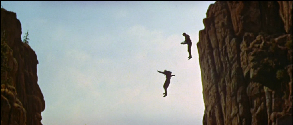 A deep gorge separates two towering rock walls under a bright daylight sky. Two men are captured in mid-air as they fall between the cliffs.