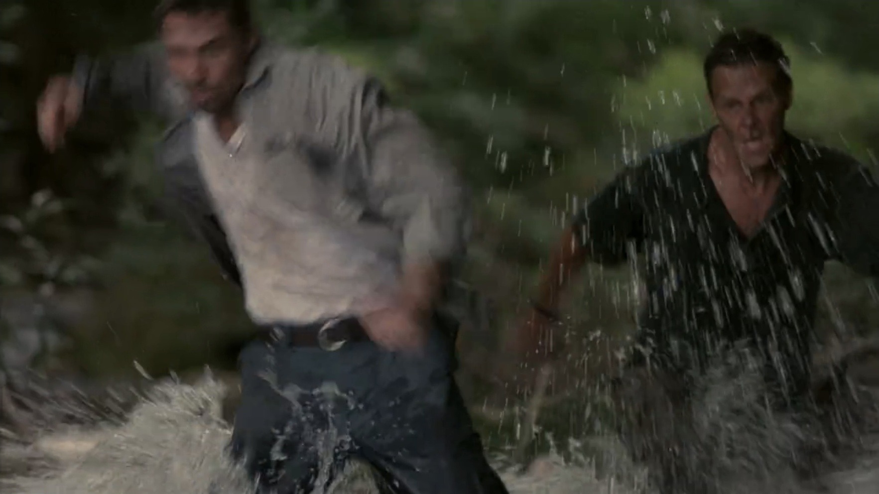 Two men are running through a shallow, dark forest river. Water sprays around their legs as they move toward the camera.