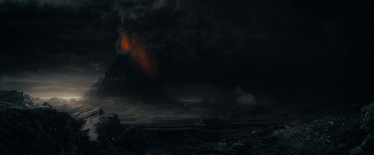 A dark, craggy landscape features a volcanic mountain obscured by heavy, thick smoke clouds. Orange magma glows from within the vent of the active, erupting mountain peak.