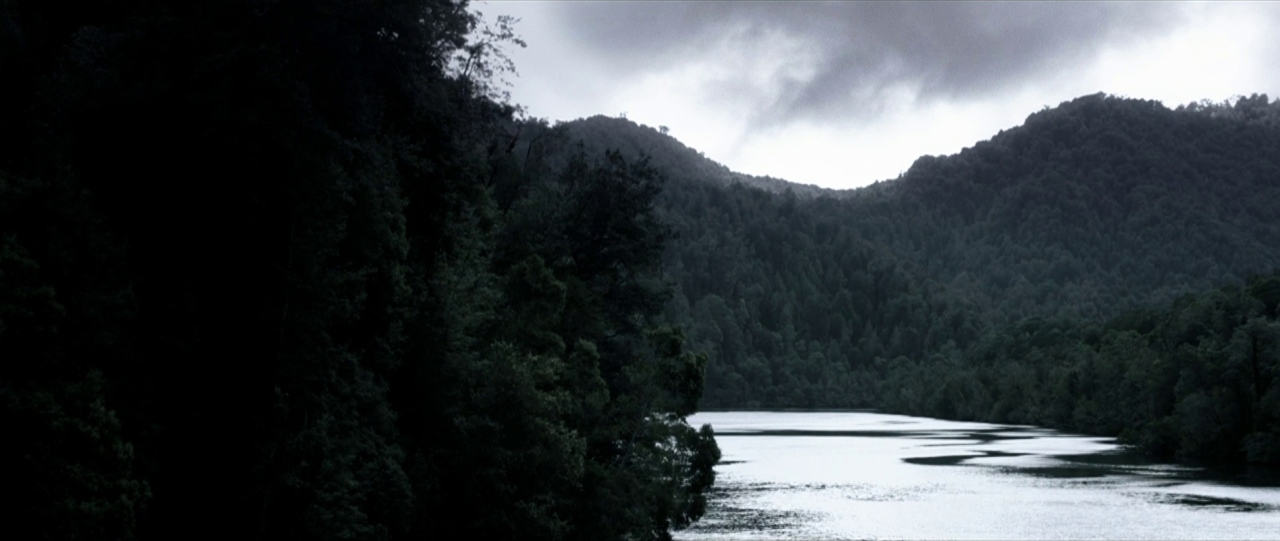 A dark river flows between dense evergreen forests under a heavy overcast sky. The river surface appears still with muted light reflections beneath tall treed hills.