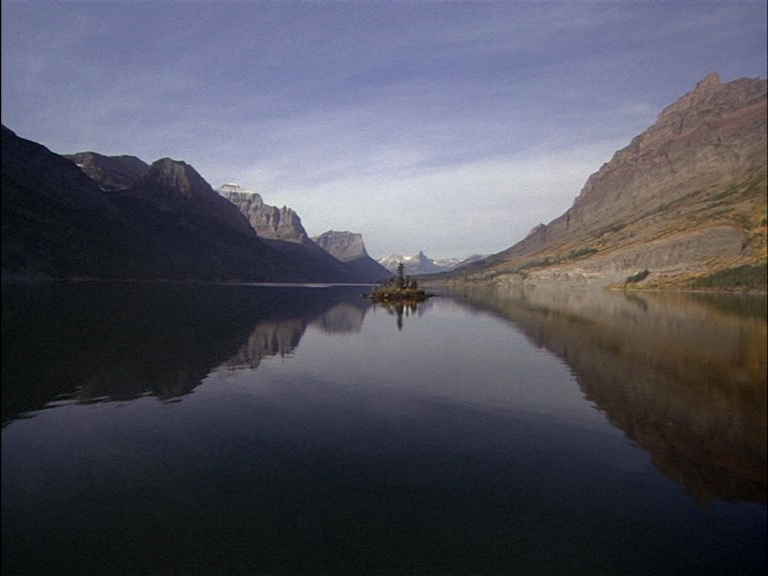 A still, reflective mountain lake sits between two large, rocky, layered mountain ranges. A small, pine-covered island rests in the center of the dark, clear water.