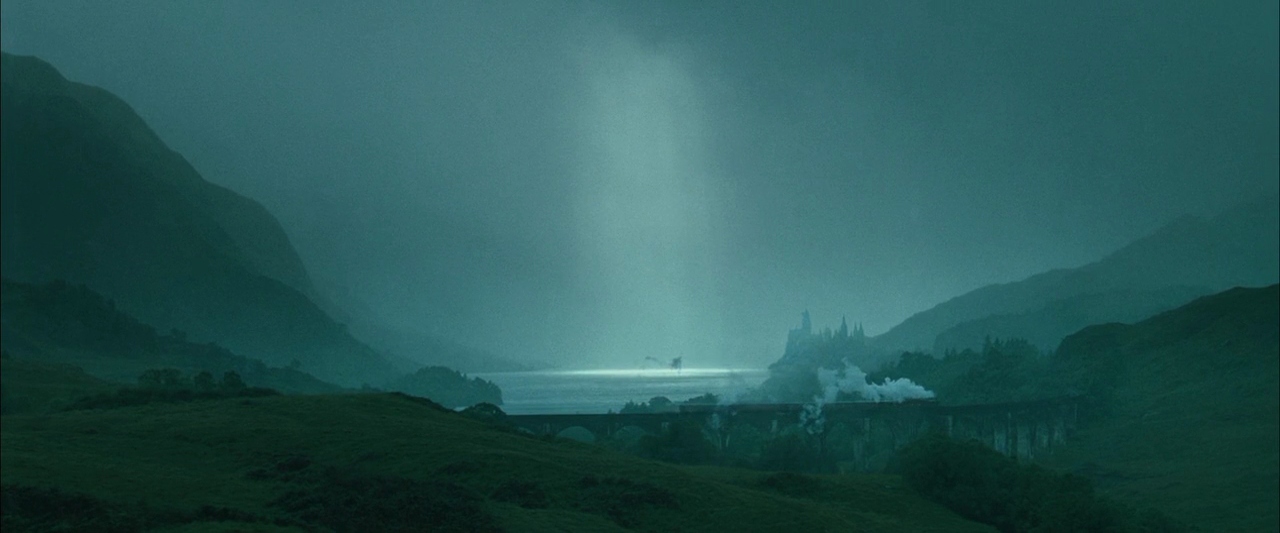 A vast mountain valley features a large lake beneath a heavy overcast sky. A steam train crosses a multi-arched stone bridge as light beams down upon the water.