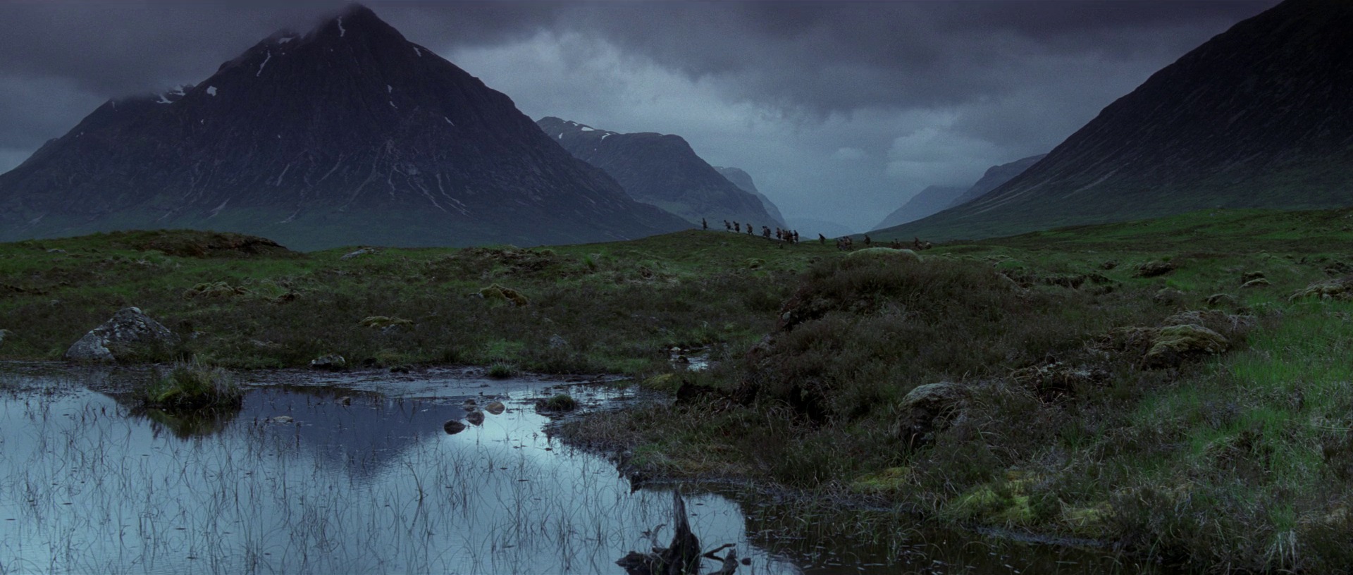 A vast, moody Scottish highland landscape features craggy green mountains under a heavy overcast sky. A small, indistinct group of people walks along a distant grassy ridge near a pool.