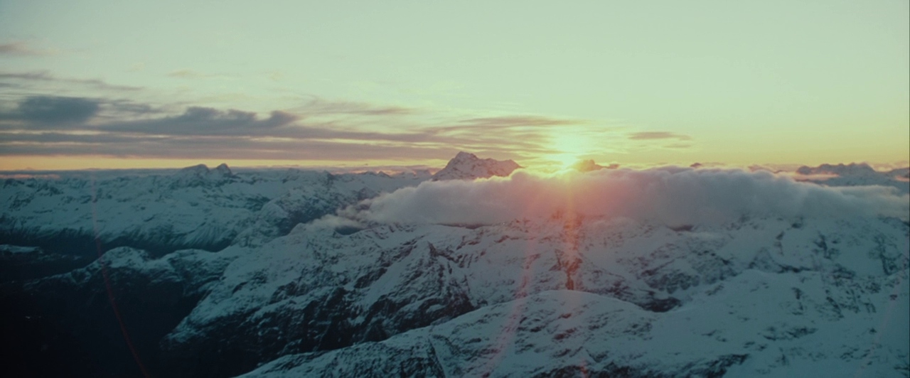 A vast mountain range is covered in deep, snow-capped peaks under a hazy sky. The sun rises over the horizon, casting light through thin cloud layers.