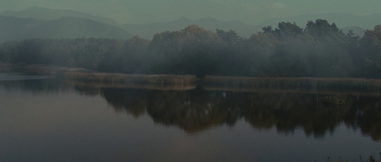 A calm lake sits in the foreground with reeds lining the shoreline. Dense forest and rolling mountains shrouded in morning mist appear in the distance.