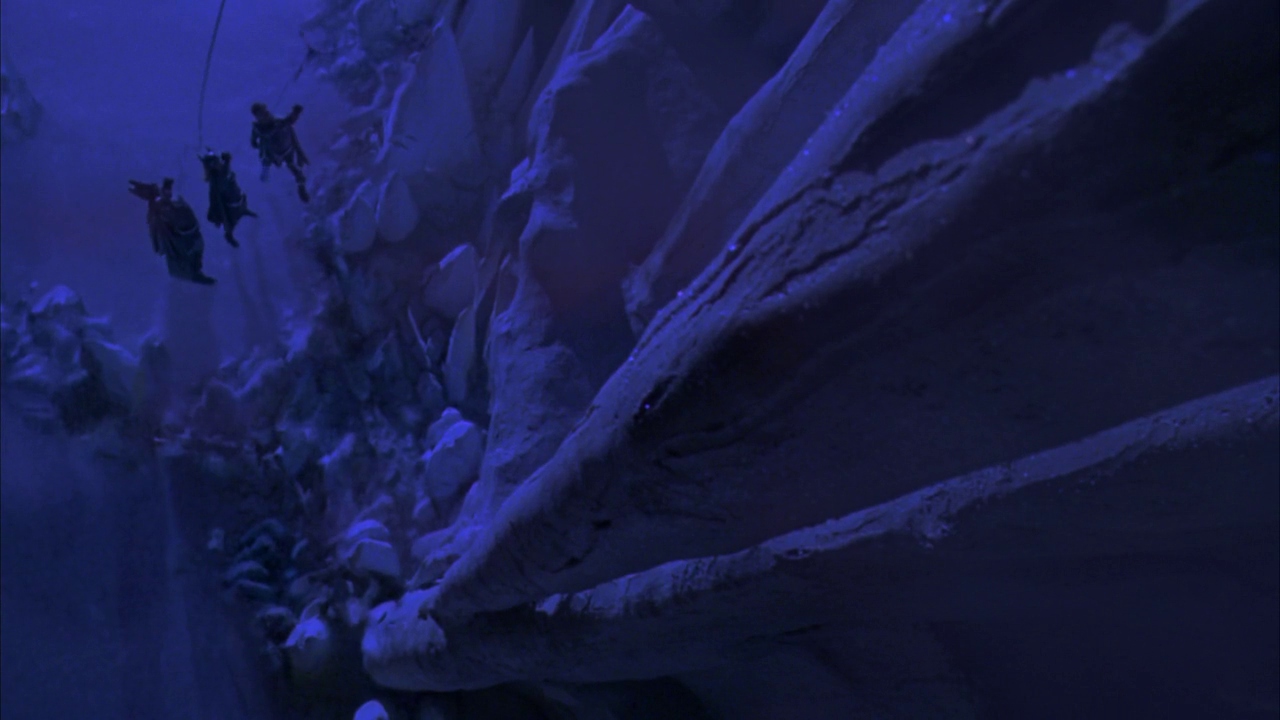 A vertical expanse of jagged blue ice walls dominates the frame. Small figures climb upward using thin ropes attached to the frozen structure.