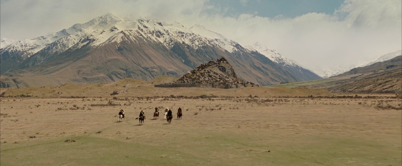 A wide, arid plain sits beneath a massive, snow-capped mountain range under a cloudy sky. Five riders on horseback travel across the dry grass toward a stone-walled ruin.