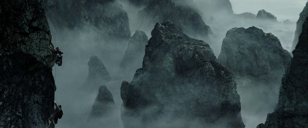 Massive jagged rock pinnacles rise through a thick layer of grey mountain mist. Two small human figures climb the vertical face of a steep cliffside.
