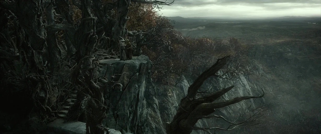 A high rocky cliffside overlooks a sprawling valley under a heavy, overcast sky. Two robed figures stand near the edge of the cliff among twisted, leafless trees.