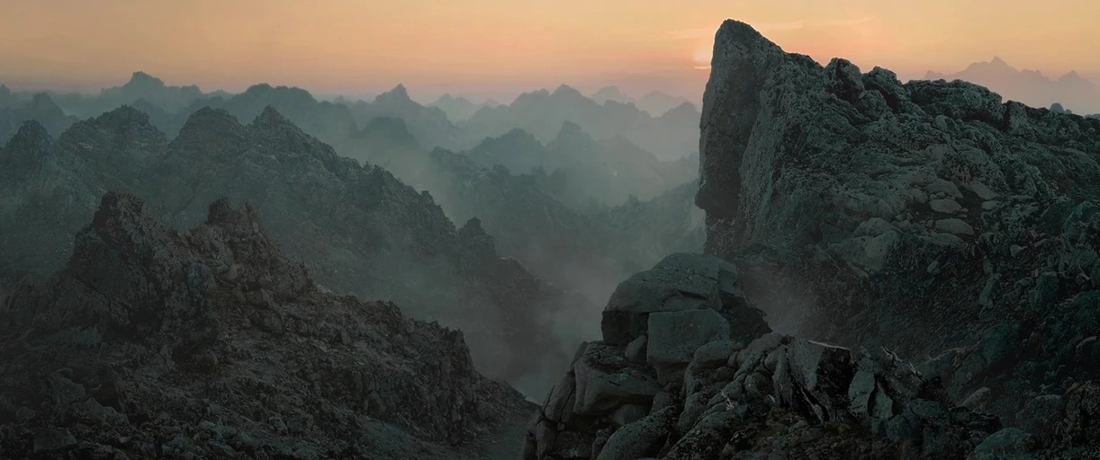A vast expanse of jagged, rocky mountain peaks stretches toward a hazy distant horizon. Thin patches of low-lying fog drift between the shadowy crevices during a hazy sunrise.
