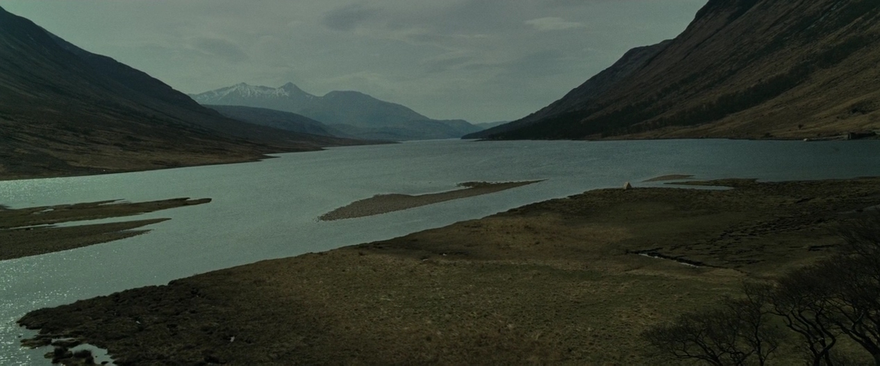 A vast loch sits between tall grass-covered mountains under a heavy overcast sky. Small gravel islands emerge from the water with snow visible on the distant peaks.