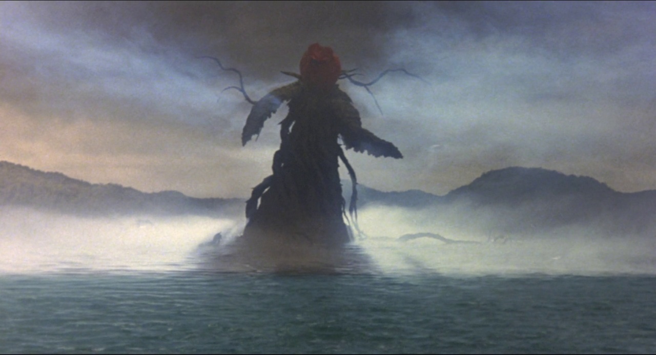 A dark, twisting, plant-like entity emerges from the center of a foggy lake. Distant hills rise from the water on both sides under a clouded sky.