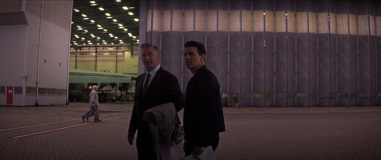 A massive industrial aircraft hangar features sliding accordion metal doors and concrete flooring. Two men walk across the tarmac in the foreground while a third figure walks behind.