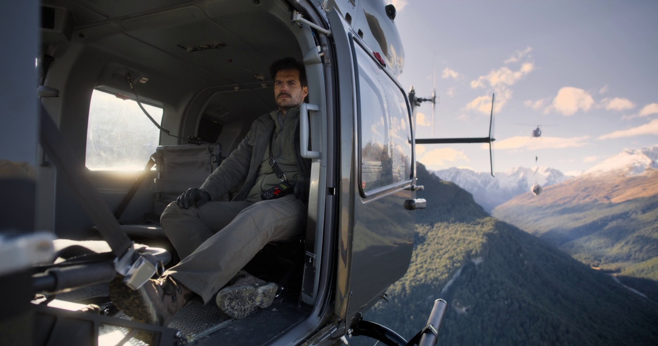 The interior of a helicopter cabin with an open door looking out over mountains. A man sits in the passenger seat wearing tactical gear and looking ahead.