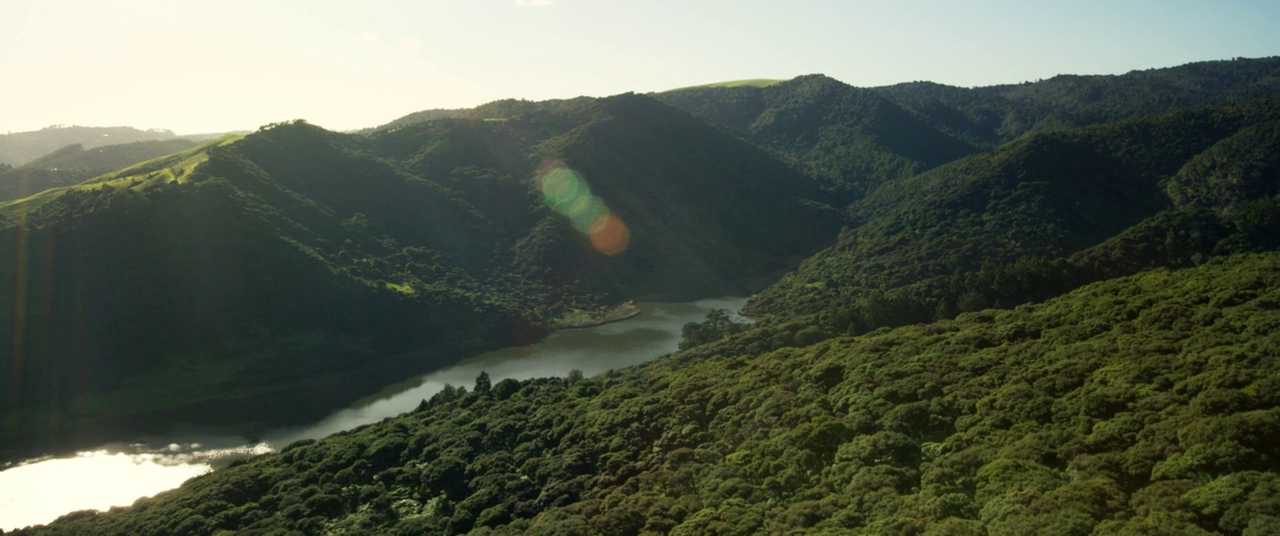 A river flows through a dense, forested valley surrounded by undulating green hills. Sunlight reflects off the water surface as a lens flare appears over the ridge.