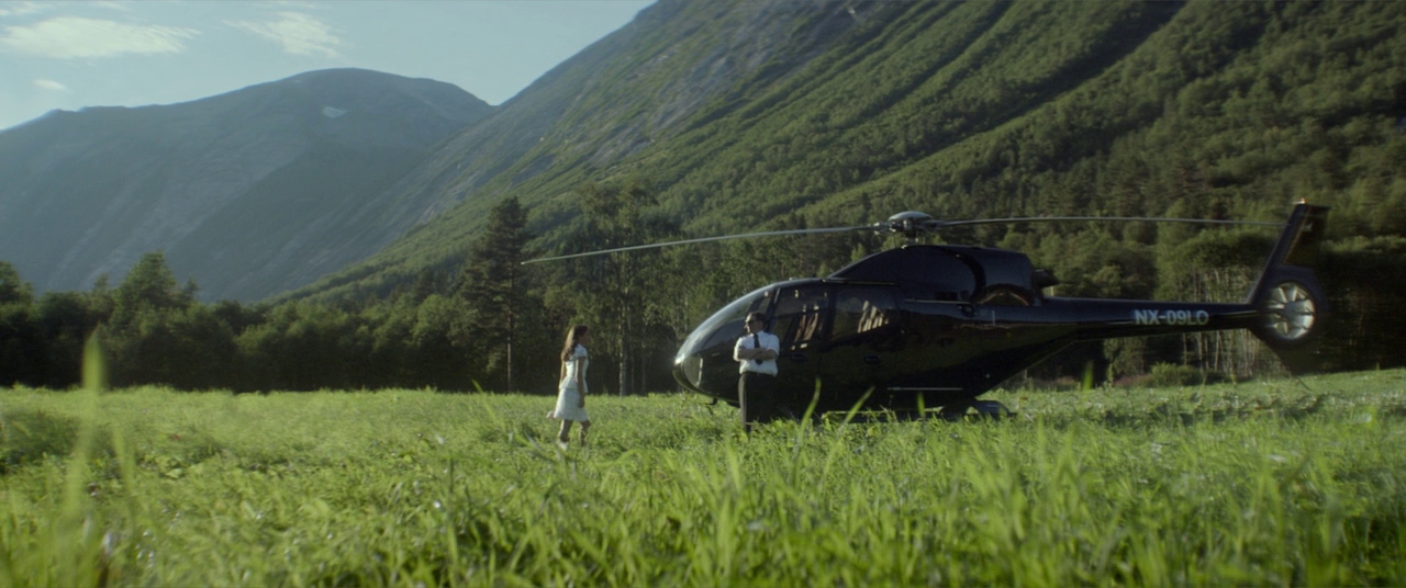 A green mountain meadow with a tall forested ridge rising in the background. A black helicopter sits on the grass while two individuals stand near the fuselage.
