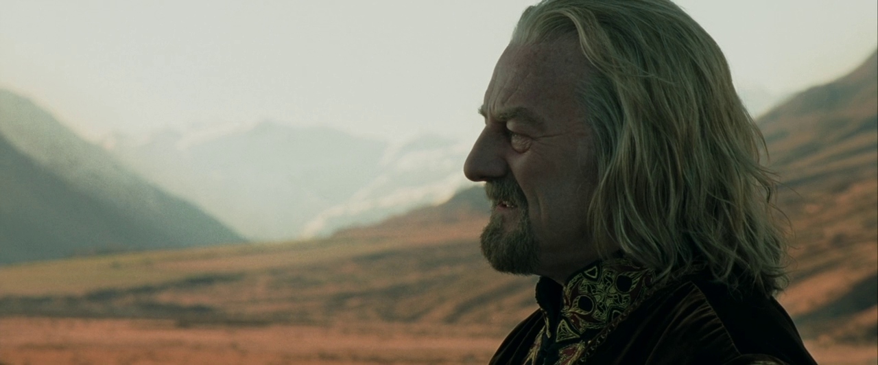 A man stands outdoors in a vast valley surrounded by distant snow-capped mountain peaks. He keeps his head in profile while gazing across the open landscape.