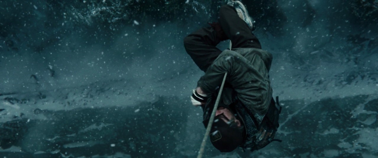 A climber descends a taut rope against a backdrop of steep, snowy mountain slopes. Heavy snow fills the air as the climber hangs inverted while managing their gear.