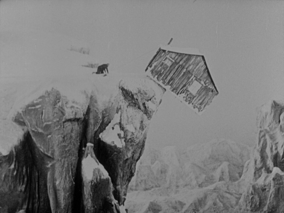 A wooden cabin balances precariously on the edge of a jagged, snow-covered cliffside. A lone person crawls through deep snow on the ridge toward the cabin.