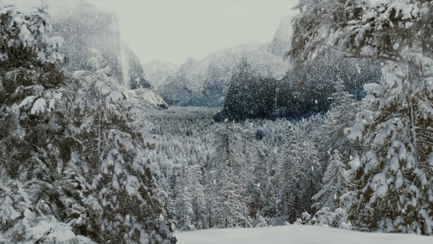 A vast snowy mountain valley is surrounded by high rocky cliffs and dense forests. Snowflakes fill the air as they descend upon the frozen trees and ground.
