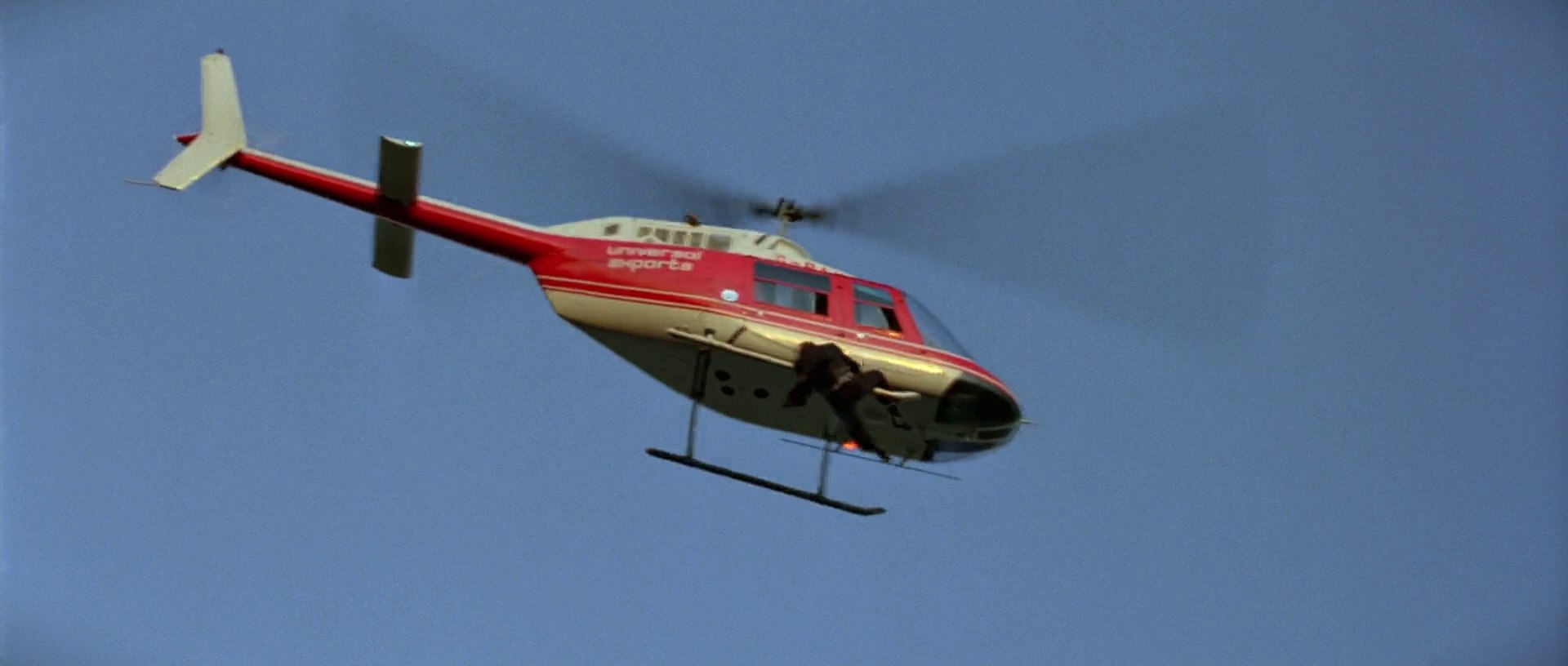 A red and white helicopter flies through a clear blue sky. A man hangs from the skid of the moving aircraft.