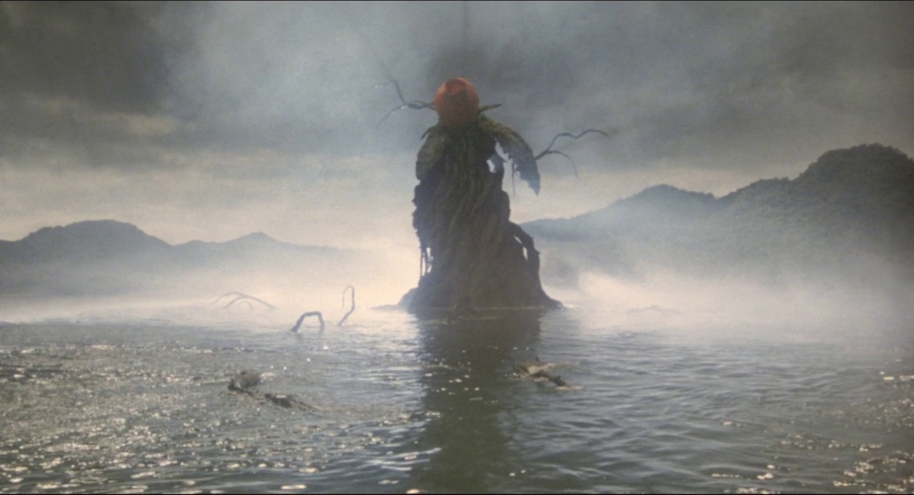 A dark, plant-like entity rises from a calm lake surrounded by misty hills. The environment is shrouded in fog under an overcast sky.