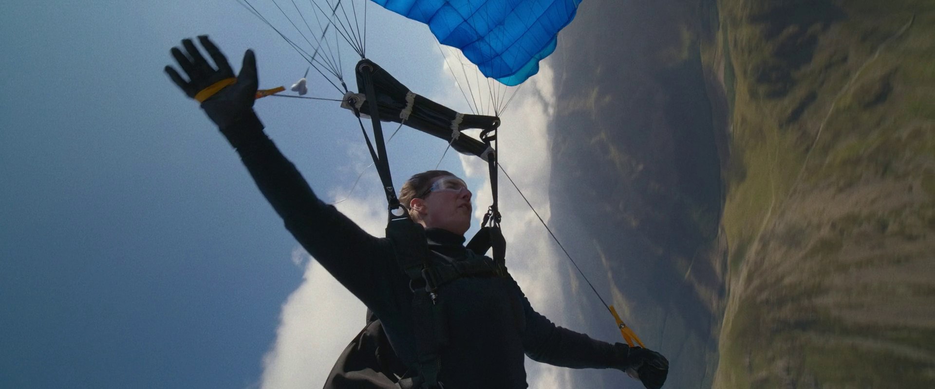 A person floats through the air using a blue parachute over mountainous landscape. The subject hangs from the harness with arms extended while wearing protective goggles.