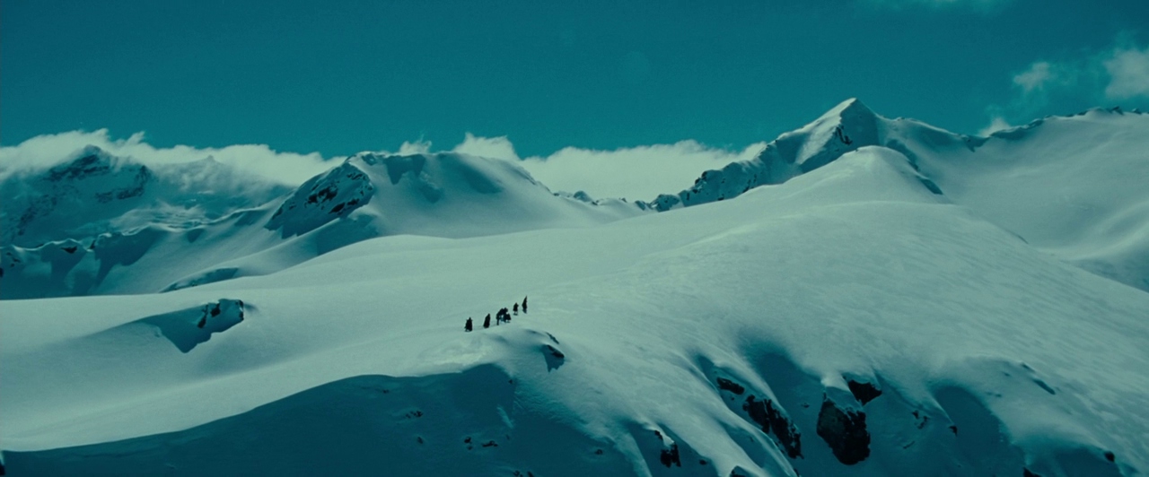 Massive snow-covered mountain peaks stretch across the frame under a clear, bright sky. A tiny group of travelers moves across a windswept, icy ridge in the distance.