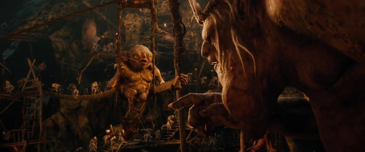 A dark subterranean cavern filled with wooden scaffolds and interconnected walkways. A giant goblin points toward a smaller goblin figure suspended in a wooden hanging cage.