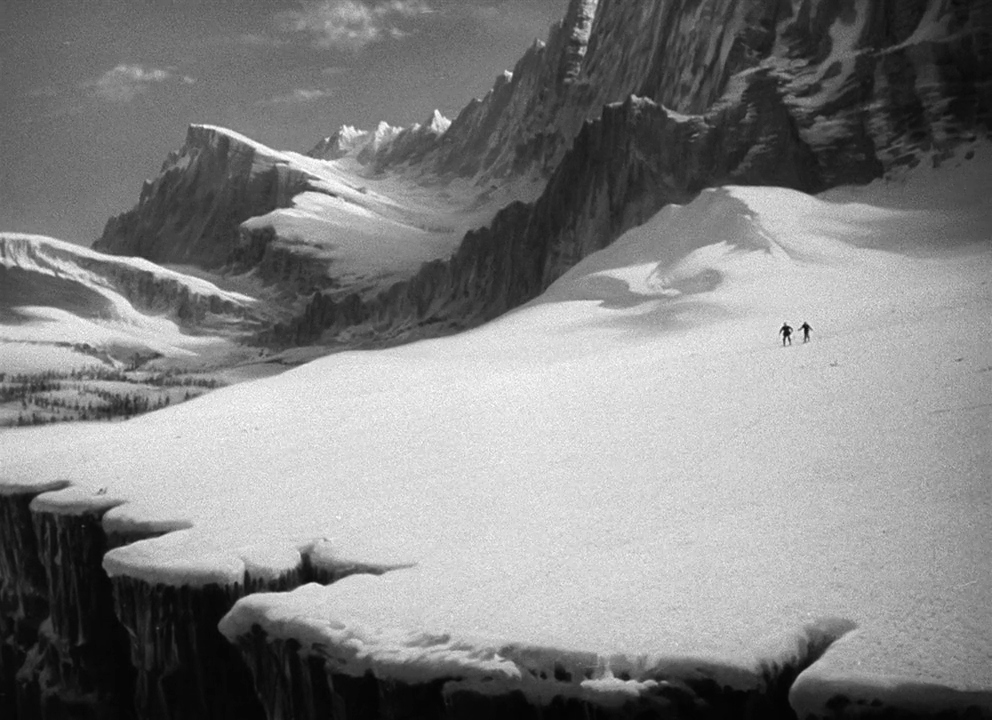 A vast, frozen high-altitude mountain range dominates the frame with craggy snow-covered peaks. Two tiny figures walk across the expansive, bright white snowy plateau towards the mountains.