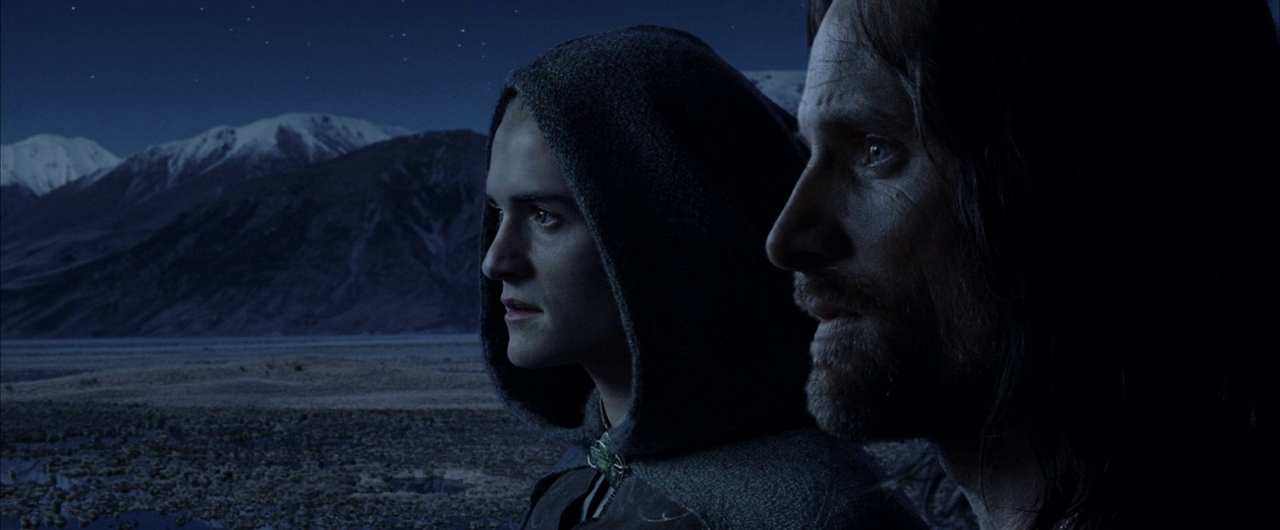 Two men stand together at night looking out across a vast, barren mountain range. The starlit sky is clear and the snowy peaks are visible in the distance.