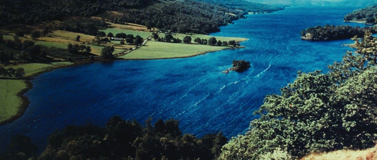 A wide aerial view of a deep blue lake surrounded by rolling green hills. Dense deciduous trees frame the foreground while pastures stretch along the winding shoreline.
