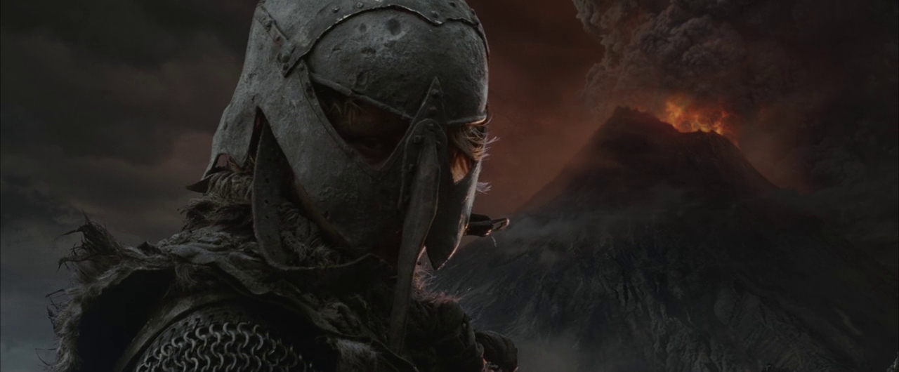A rugged volcanic landscape at night features an active, glowing mountain peak in the distance. A soldier in weathered iron armor looks forward while ash clouds fill the dark sky.
