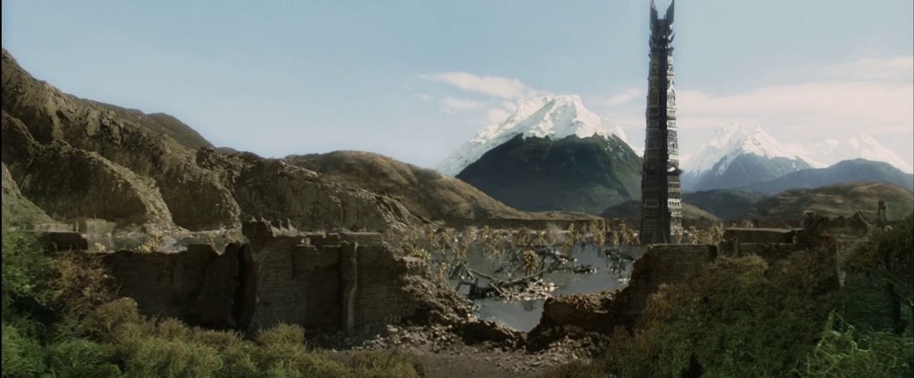 A vast, ruined stone fortification sits in a valley under a clear blue sky. A tall, dark, spired tower stands prominently in the center against snow-capped mountain peaks.