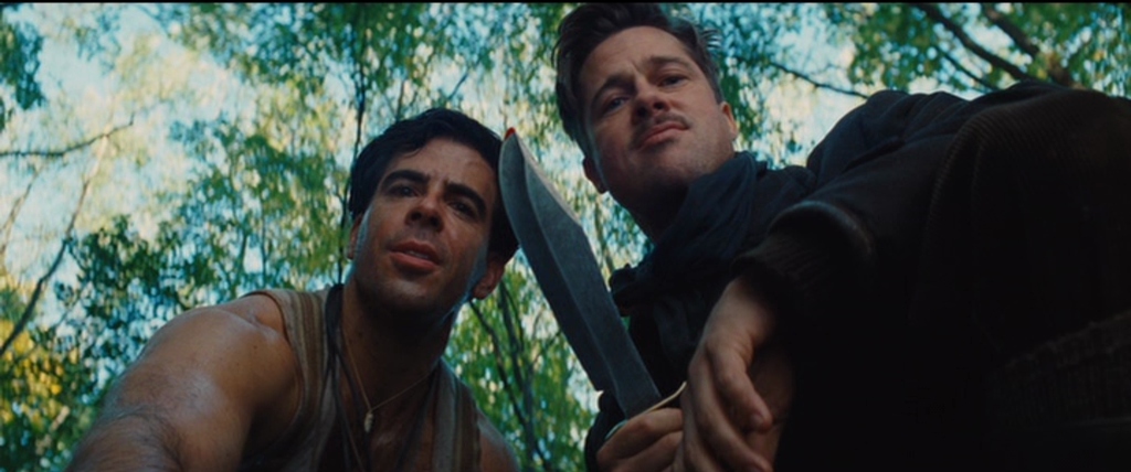 A dense green woodland setting with sunlight filtering through the trees. Two men in period military-style clothing look downward toward the ground.