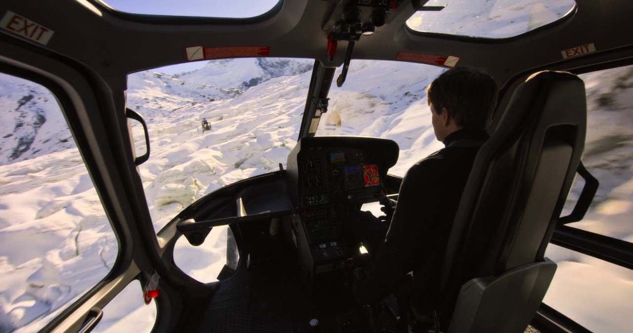 The interior of a helicopter cockpit displays modern flight instruments and large observation windows. The pilot controls the aircraft as it flies over a vast expanse of snow.