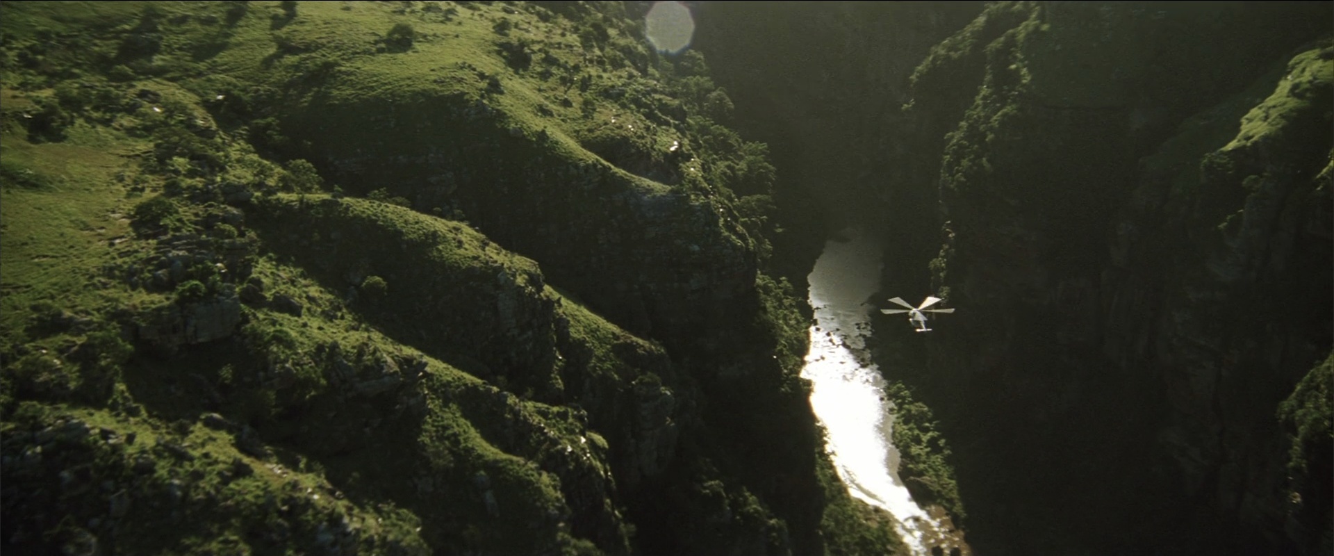 A deep rocky river canyon covered in green grass and steep cliffs. A helicopter flies over the narrow river at the base of the gorge.