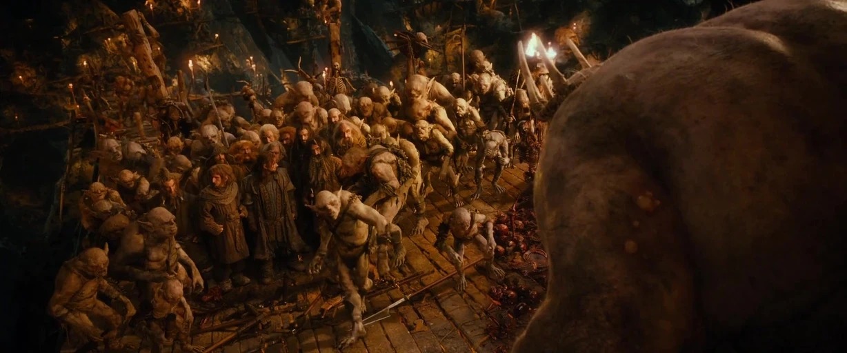 A subterranean cavern is packed with a dense crowd of goblins and captives. The creatures stand on wooden platforms with torches mounted to structural pillars.