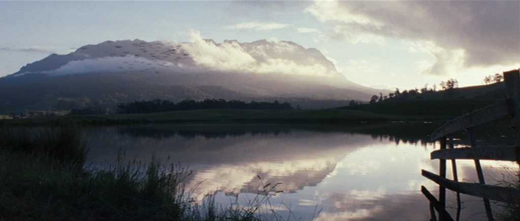 A wide mountain range sits behind a calm lake under a cloudy sky. A wooden fence stands in the foreground to the right near the water edge.
