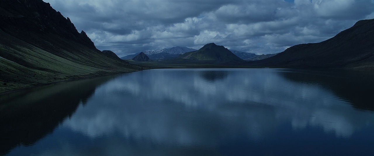 A vast alpine lake sits surrounded by steep, green-sloped mountains under a heavy overcast sky. The water reflects the dark clouds and surrounding terrain in its calm, glassy surface.