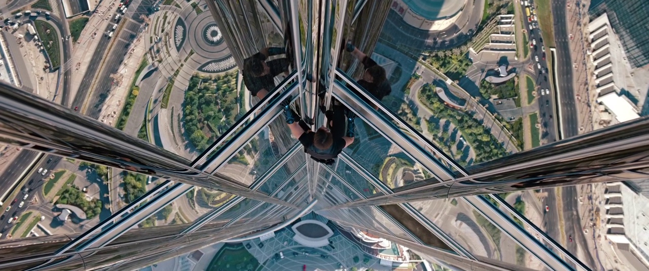 A man climbs the exterior glass facade of a towering skyscraper from a high perspective. The city infrastructure and road networks are visible far below the climber.