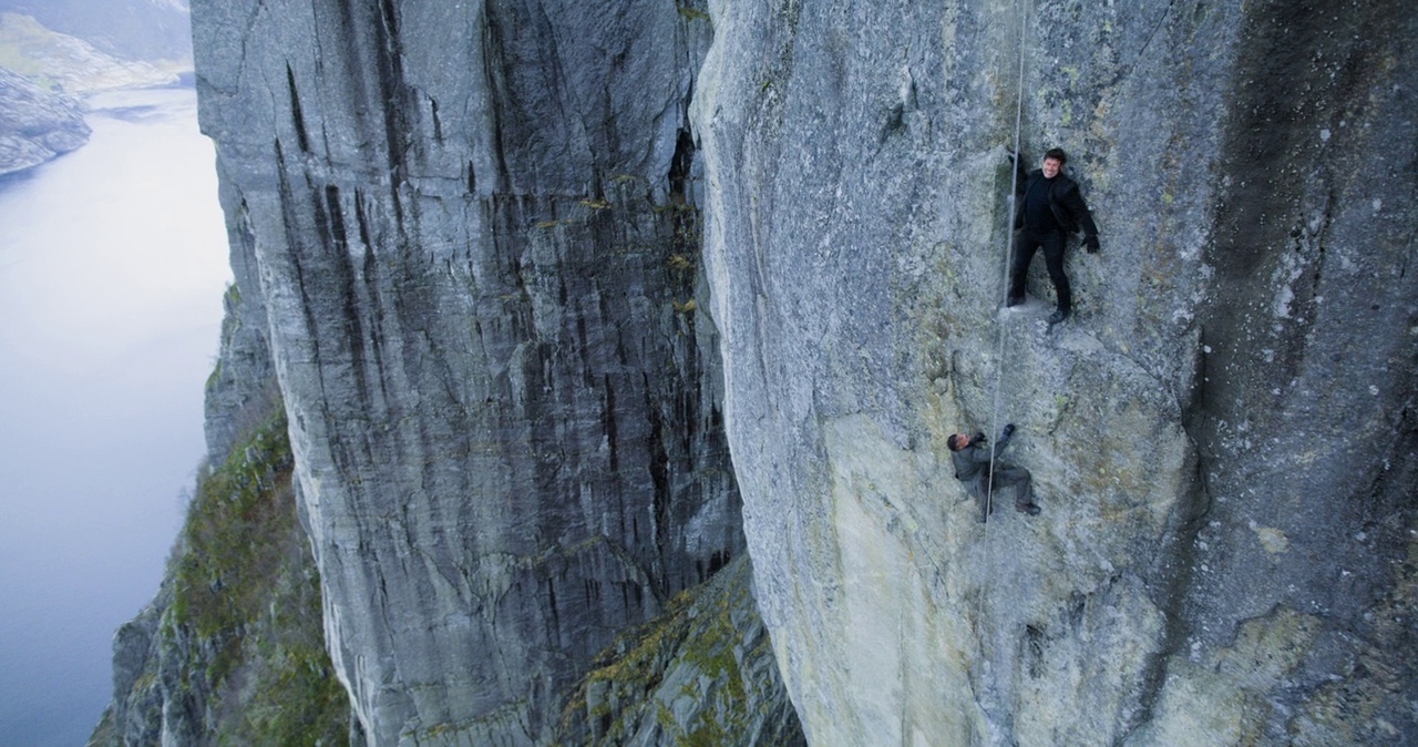 A massive vertical cliff wall rises above a deep blue body of water. Two men are anchored to the rock face by a single vertical rope.