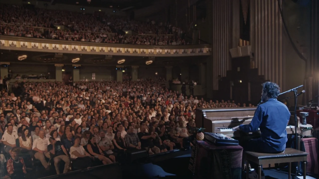 practical lighting example from Flight of the Conchords: Live in London: A dark, multi-level concert hall is filled with a large seated audience. A ma