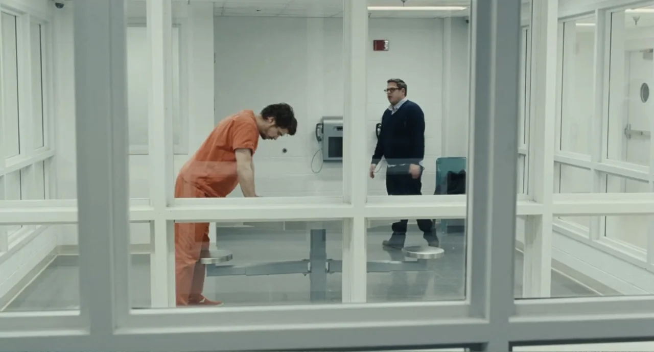 deep focus example from Bad Boys: A True Story: A stark white visitation room has a metal table and white tiled walls. A man in an orange jumpsuit lea
