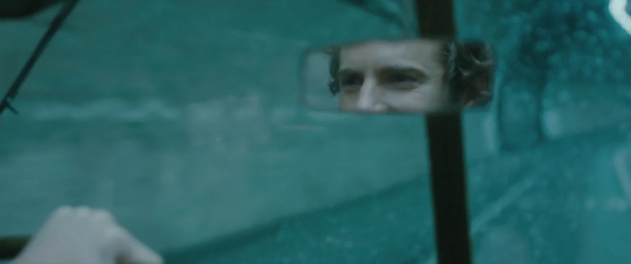 window light example from Ginger & Rosa: A view from inside a moving vehicle through the tinted front windshield. A man's face is visible within the r